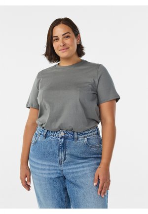 Donna con capelli corti e scuri che indossa una maglietta grigia infilata in jeans blu a vita alta, in piedi davanti a uno sfondo bianco uniforme.