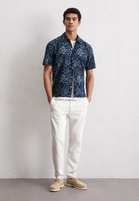Korte mouwen marineblauw shirt met een wit bloemenpatroon, gecombineerd met witte broeken en beige schoenen. Het shirt heeft een knoopsluiting aan de voorkant en een kraag.