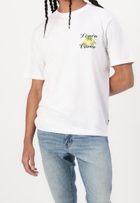 Valkoinen puuvillapaita lyhyillä hihoilla, jossa on pieni vihreä ja keltainen sitrushedelmäkuvio ja teksti "Limón Citrico" rintamirkkissä.