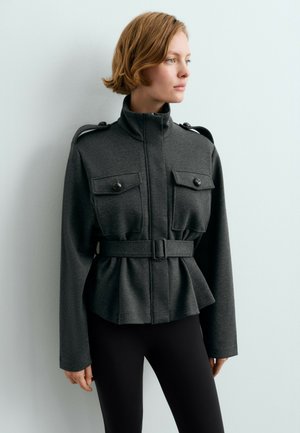 Femme portant une veste ceinturée gris foncé avec un col montant et des poches poitrine boutonnées, debout contre un mur gris clair, regardant vers la droite.