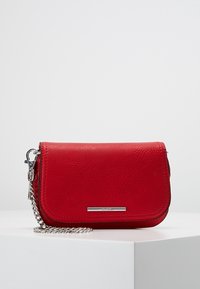 Pochette en cuir rouge avec bandoulière chaîne argentée et petite plaque métallique rectangulaire à logo sur le devant, exposée sur un socle blanc.