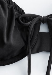 Gros plan d'un tissu noir avec un bord élastique froncé et une boucle de cordon nouée, mettant en valeur la texture et les détails de couture.
