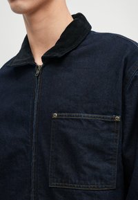 Tamsiai mėlynas džinsinis švarkas su aksominiu apykakle, priekinis užtrauktukas ir pleistrinė kišenė. Siūlėse yra kontrastingos geltonos siūlės.