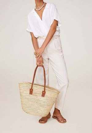 Grote geweven stro tote bag met gevlochten bruine handvatten en bruine leren accenten. Model draagt een witte blouse en crèmekleurige broek.