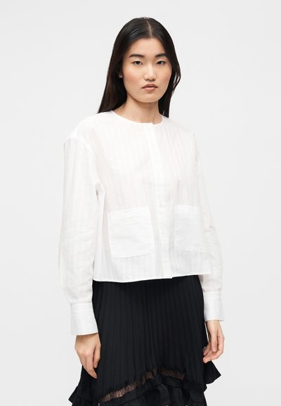 Άσπρη κοντή μπλούζα με κάθετες ρίγες, στρογγυλή λαιμόκοψη και μακριά μανίκια με δύο τσέπες μπροστά. Το ύφασμα φαίνεται ελαφρύ και υφασμένο.