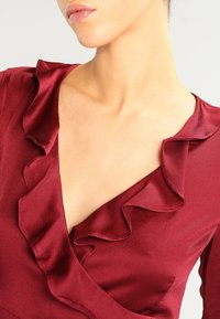 Top en satin bourgogne avec un décolleté en V profond et des détails de volants le long des bords, texture lisse et silhouette ajustée.