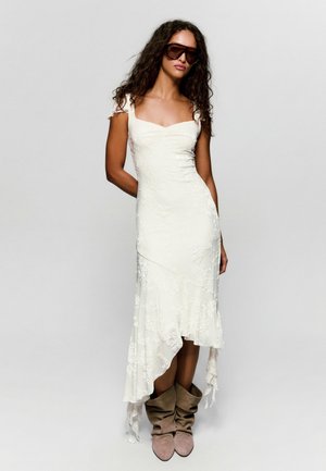 Nő, hosszú göndör hajjal, sötét napszemüveget visel, fehér aszimmetrikus csipkeruhában és lezser bézs csizmában, egyszínű háttér előtt áll.