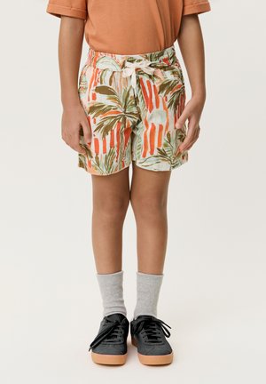 Enfant portant un short orange à imprimé palmiers, des chaussettes gris clair, des baskets noires avec semelles en gomme et un T-shirt orange à manches retroussées.