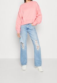 Sudadera rosa corta con escote redondo y logo bordado; jeans de pierna ancha azul claro con detalles desgastados; zapatillas blancas.