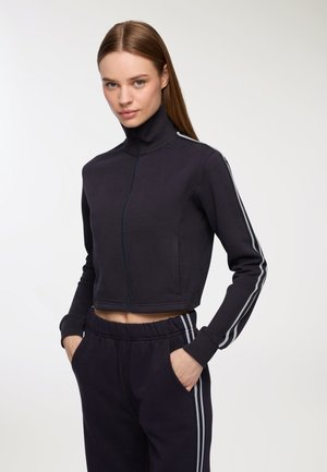 Giovane donna che indossa una giacca a zip accorciata di un blu navy scuro e pantaloni da tuta abbinati con strisce bianche sui lati, mani nelle tasche, guarda verso la macchina fotografica.