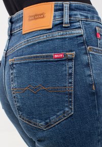 Tummansinisiä farkkuja, joissa on nahkainen "BIG STAR" -merkki vyötäröllä, punainen lappu takataskussa ja koristeellinen ompelu.