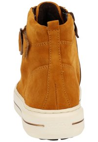 Botín de ante en color mostaza con una suela de goma blanca. Presenta un cierre de cremallera y un botón decorativo en el costado. Acabado texturizado.