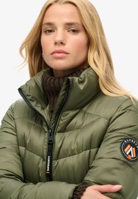 Oliwkowa kurtka puchowa z wysokim kołnierzem, zapinana na zamek z widocznymi szwami. Posiada okrągłą naszywkę z logo na lewym rękawie.