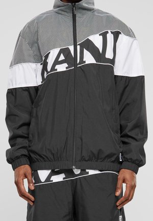 Person trägt eine schwarz-weiße Jacke mit Reißverschluss, elastischen Bündchen und fettem Schriftzug über der Brust sowie passende schwarze Hose mit weißem Schriftzug.