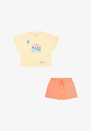 Camiseta amarilla de manga corta con el texto gráfico "MAKE WAVES" en azul, combinada con pantalones cortos coral con cintura elástica y detalle del logo.