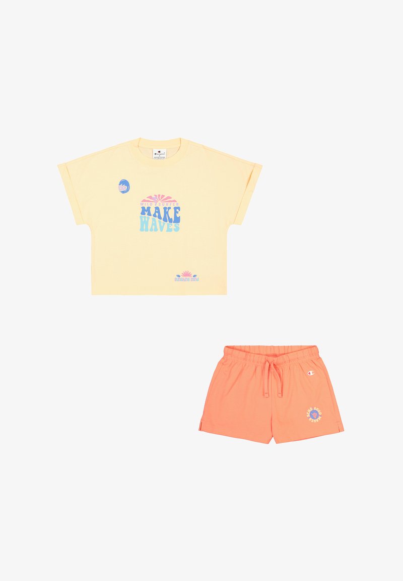 Camiseta amarilla de manga corta con el texto gráfico "MAKE WAVES" en azul, combinada con pantalones cortos coral con cintura elástica y detalle del logo.