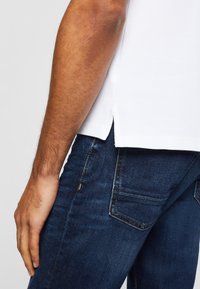 Chemise blanche à manches courtes avec fentes sur les côtés, associée à un jean en denim bleu foncé avec des coutures contrastantes et une poche de chaque côté.