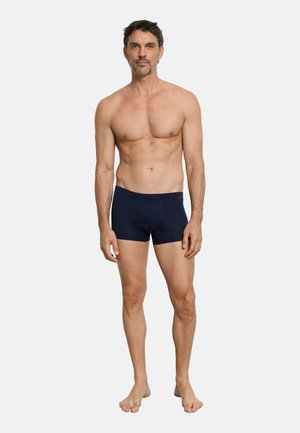 Homme adulte debout, pieds nus, portant un boxer bleu marine, sur un fond clair uni, face à l'avant avec les bras détendus.