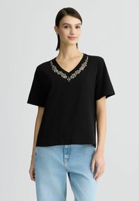 T-shirt nera a maniche corte con scollatura a V, caratterizzata da un motivo decorativo di perline attorno al collo. Realizzata in un tessuto morbido.