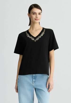 Sort kortærmet t-shirt med V-hals, der har et dekorativt perlemønster omkring halsudskæringen. Fremstillet af et glat stof.