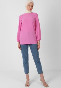 Maglietta a maniche lunghe rosa con polsini arricciati, abbinata a jeans azzurri chiari. Tessuto morbido, vestibilità rilassata e design semplice.
