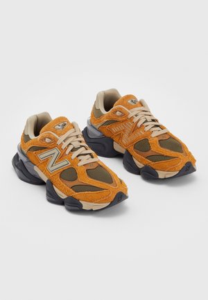 Paar orangefarbene und olivgrüne New Balance Sneaker mit beigen Schnürsenkeln und klobigen schwarz-beigen Sohlen auf weißem Hintergrund.