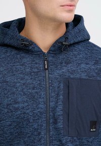 Tefto modra zip-up kapuca z teksturirano tkanino, nastavljivo vrvico in kontrastno žep z logotipom na levi strani.