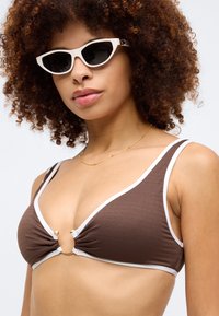 Brown ribbed bikini top with white trim, featuring a gold ring accent at the center. Paired with white cat-eye sunglasses.