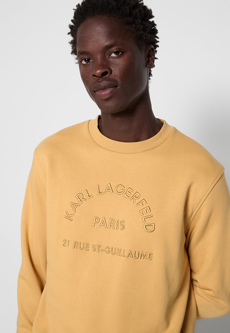 Gelbes Sweatshirt mit Rundhalsausschnitt, das auf der Vorderseite den erhabenen Text "Karl Lagerfeld Paris" und "21 Rue St-Guillaume" zeigt. Glatte Textur.