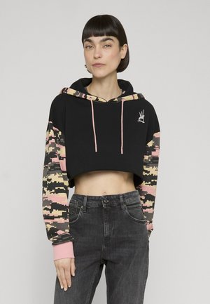 Femme portant un sweat à capuche court noir avec des manches et une doublure de capuche camouflage numérique rose et beige, associé à un jean noir taille haute.