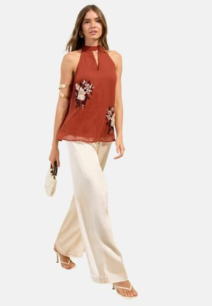 Woman walking wearing a sleeveless rust halter top with floral embroidery, cream wide-leg pants, white heels, gold arm cuff, and holding a small beige handbag.