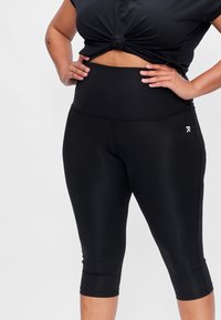 Zwarte capri leggings van rekbaar materiaal met een hoge tailleband. Bevat een klein logo-detail op het onderbeen. Glad textuur.