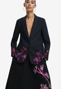 Tengerészkék blézer mély V-kivágással és rózsaszín virágos díszítéssel az ujjakon. Fekete szoknyával párosítva, amelyen illeszkedő virágminták találhatók.