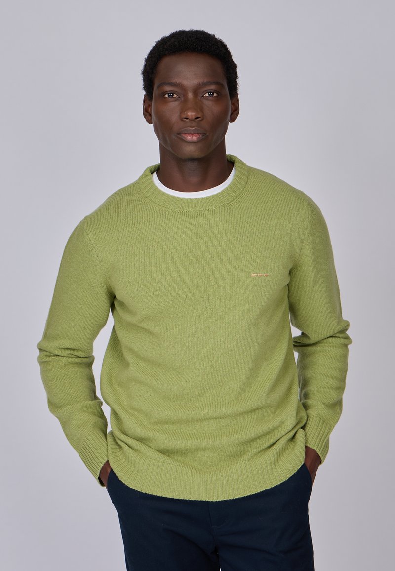 Pull tricot vert avec un col arrondi, des poignets et un ourlet côtelés, présentant un petit logo sur la poitrine. Porté par-dessus une chemise blanche.