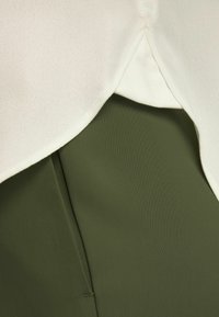 Bílá hladká látka překrývá texturovanou, olivově zelenou, pružnou tkaninu. Design má vrstvený vzhled s viditelnými švy.