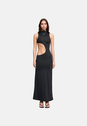 Mujer con vestido largo negro sin mangas con un gran recorte en el lado derecho, combinado con sandalias negras de punta abierta y un collar.