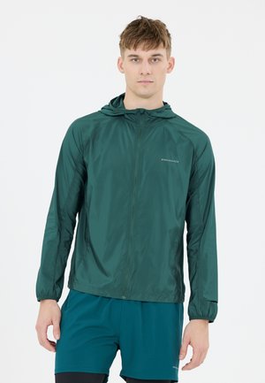 Junger Mann trägt eine dunkelgrüne, leichte Kapuzenjacke und türkisfarbene Sportsshorts, steht vor einem einfarbigen weißen Hintergrund.