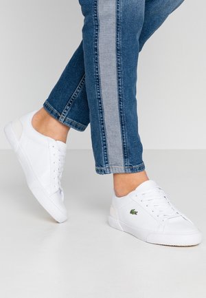 Person iført blå jeans med en grå stribe og hvide Lacoste-sneakers, det ene ben løftet, stående på en glat hvid gulv.