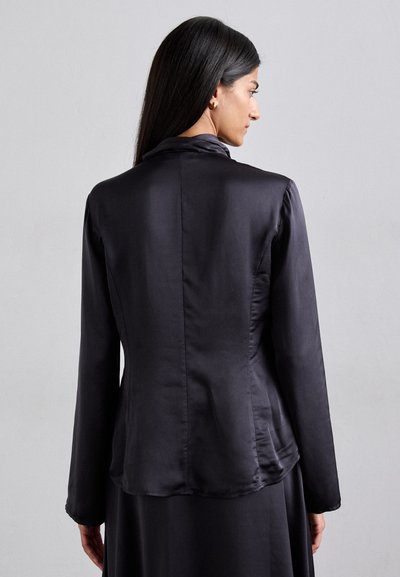 Giacca nera in satin con un design aderente, maniche lunghe e colletto a punta. Texture liscia e leggero lucido. Dettaglio della cucitura centrale classica.