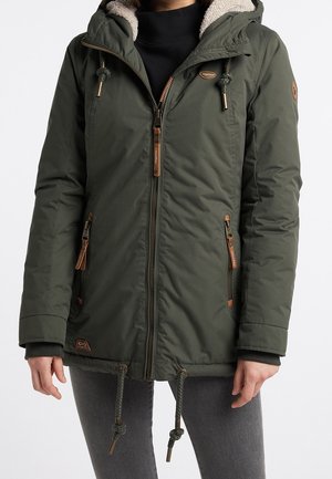 Mujer con chaqueta de invierno con capucha de color verde oscuro, cierre de cremallera y cordones, sobre una camiseta negra y pantalones grises.