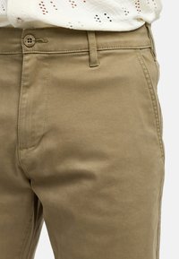 Beige khaki-housut, joissa on tasainen etuosa, sivutaskut ja tavallinen nappisulku, valmistettu sileästä ja keveästä kankaasta.