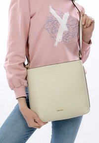 Borsa a tracolla in pelle crema con una superficie texture, caratterizzata da un logo dorato e una lunga tracolla regolabile.