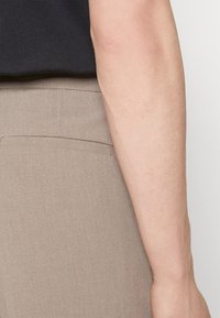 Kalhoty ze světle hnědé látky s elegantním, na míru střiženým designem; mají jedinou zadní kapsu a hladkou texturu.