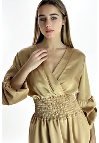 Abito in satin beige con profondo scollo a V, maniche a palloncino e vita arricciata. La texture liscia e il design fluido esaltano la silhouette del capo.