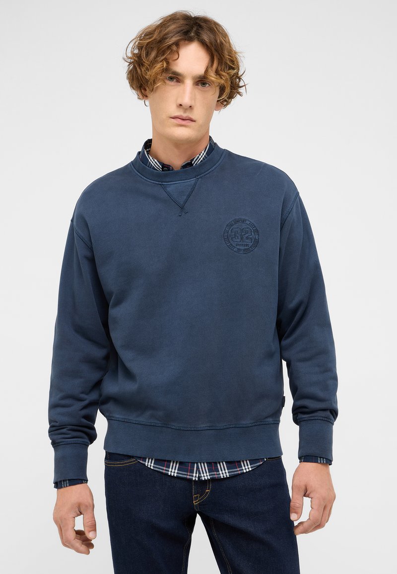 Sweat-shirt bleu marine à manches longues, poignets côtelés et col rond. Présente un logo circulaire texturé sur la poitrine. Porté sur une chemise à carreaux.