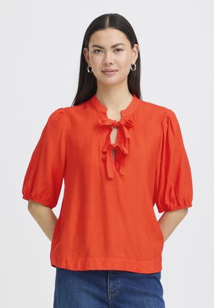 Femme aux longs cheveux foncés portant un chemisier orange vif à manches bouffantes et deux nœuds noués au niveau du col, associé à un jean bleu.