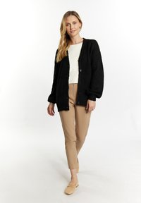 Zwarte, gemêleerde cardigan met knopen, gedragen over een crèmekleurig topje en beige broek. De cardigan heeft een ruime pasvorm en ribbels aan de mouwen.
