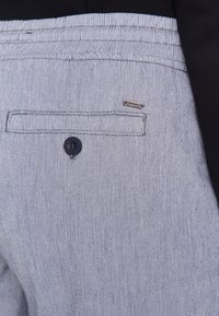 Pantalons rayés avec une poche arrière dotée d'un bouton noir et d'une étiquette en métal. Le tissu semble texturé et léger.