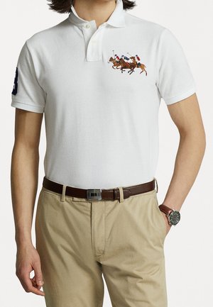 Polo shirt blanc à manches courtes, col côtelé et scène de polo brodée. Associé à un pantalon beige et une ceinture marron.