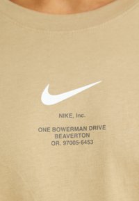 Gaišbeige T-krekls ar apaļu kakla izgriezumu, kura priekšpusē ir baltā Nike swoosh logotipa un melnā teksta ar zīmola un adreses uzdruka.